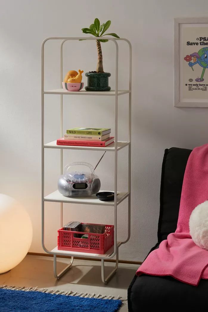 White metal Alana narrow storage shelf with four shelves, displaying a plant, books, a radio, and a storage basket. Modern shelving unit for home organization.
