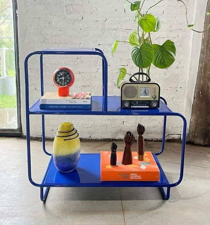Blue metal Alana Nightstand with two shelves, displaying a clock, radio, vase, and figurines. Modern shelving unit.