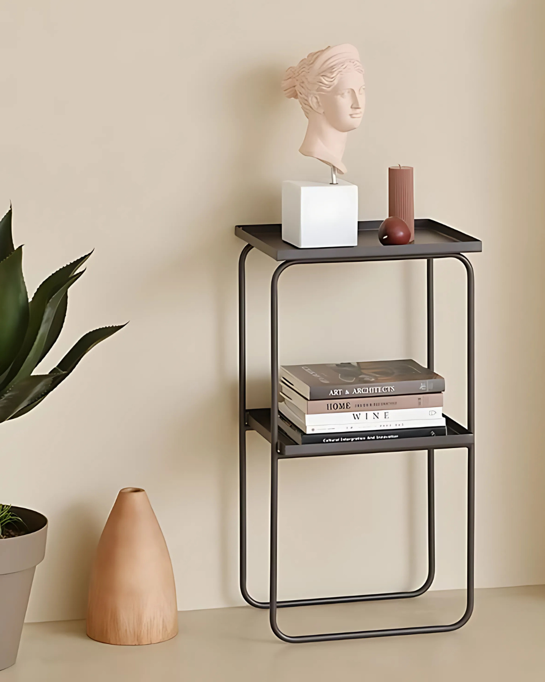 A slender black two-tiered side table displays a small white bust, a stack of books, and a brown candle on its top shelf. Below, the open lower shelf provides additional space, while a tall potted plant and a light brown vase sit beside it on the floor.