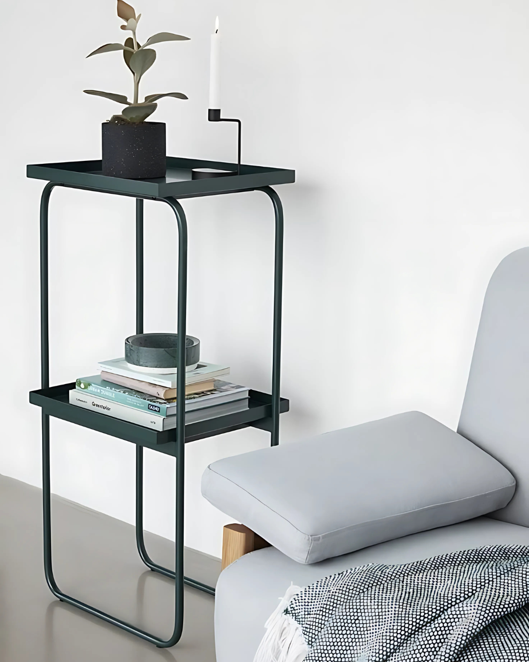 A slender black two-tiered side table displays a small white bust, a stack of books, and a brown candle on its top shelf. Below, the open lower shelf provides additional space, while a tall potted plant and a light brown vase sit beside it on the floor.