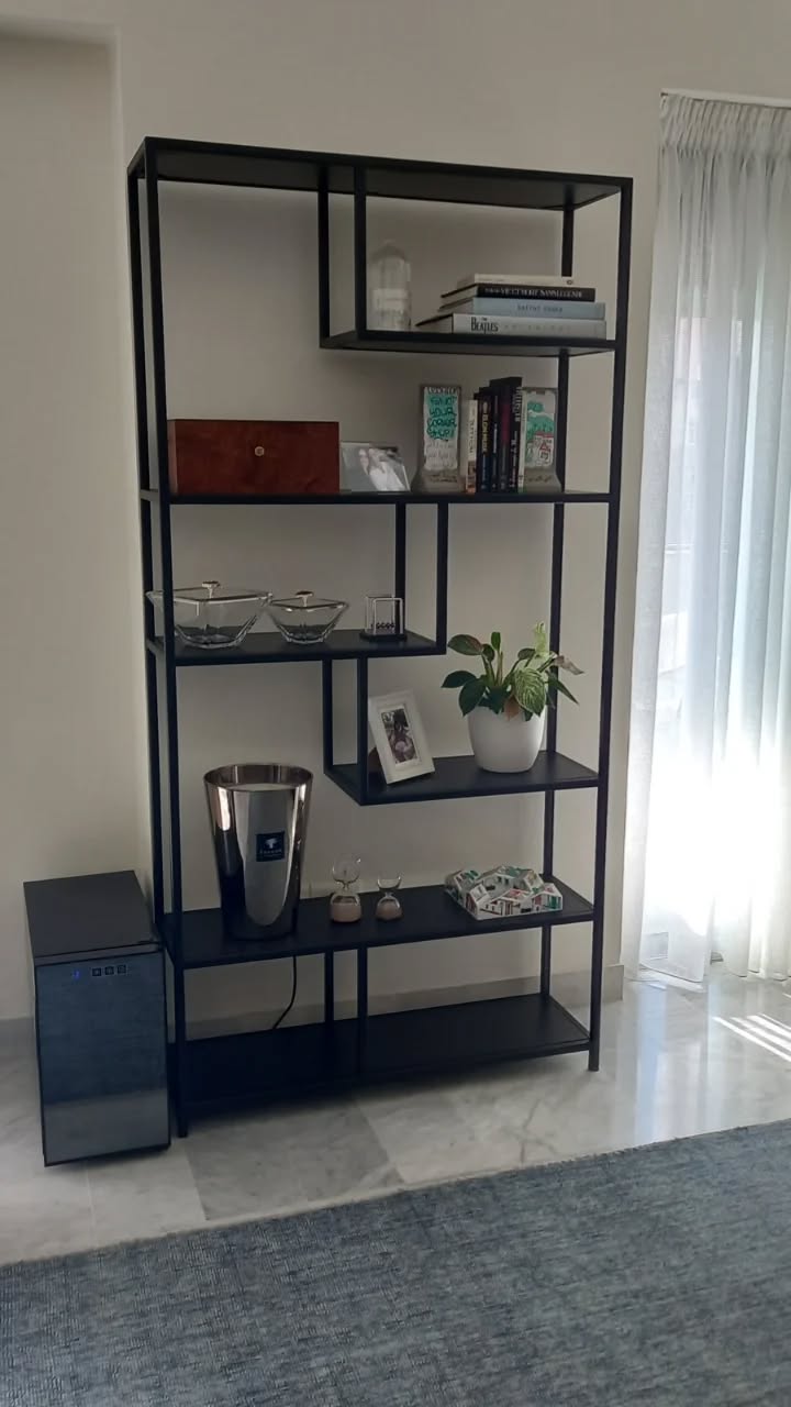 Modern black metal bookshelf with open, asymmetrical shelving. Displaying decorative plates, vases, plants, and books. Contemporary design for home or office storage and display.