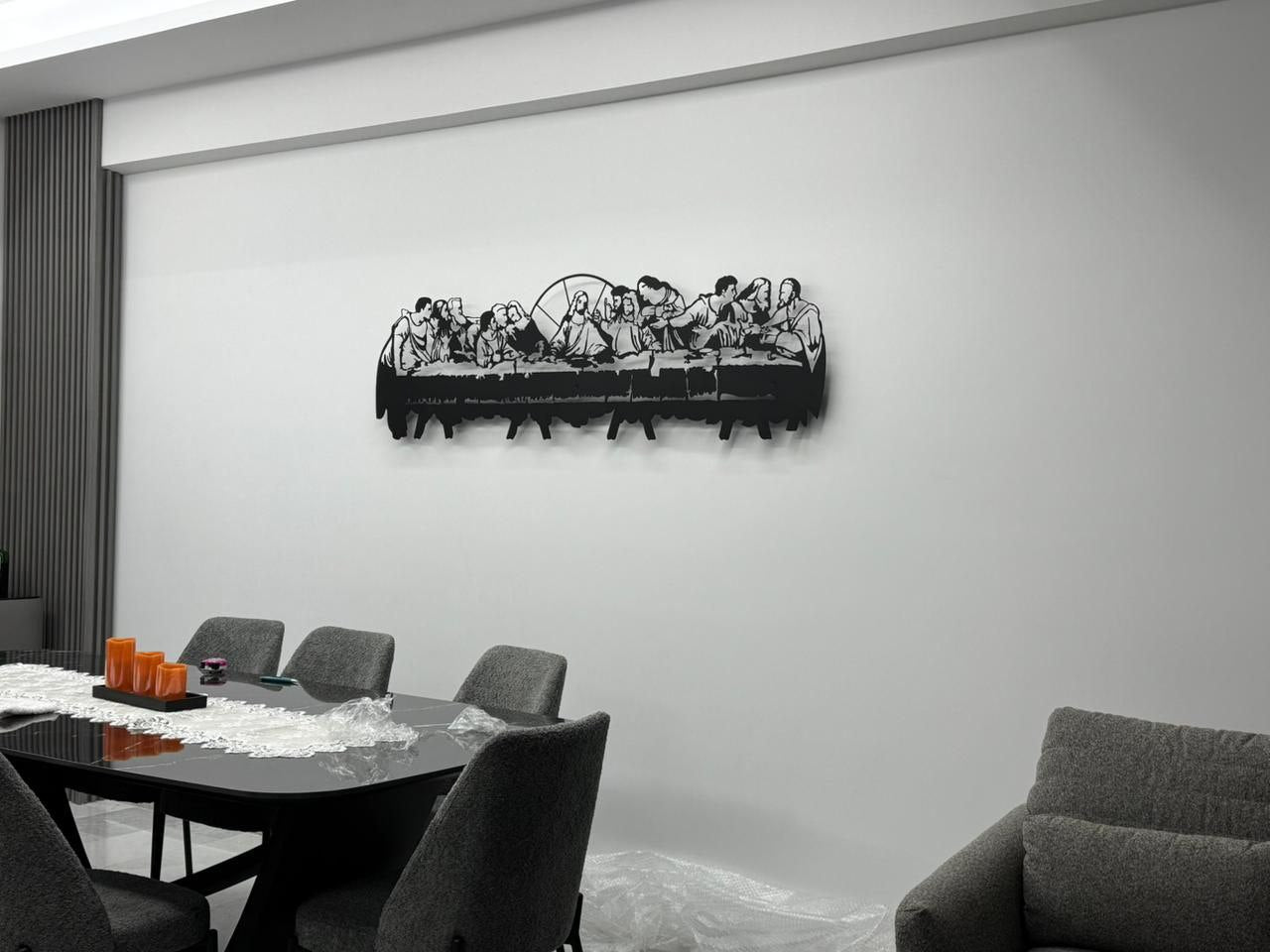 A detailed black metal silhouette of the Last Supper, showing Jesus and his disciples at a long table, mounted on a white wall. The artwork is cut out in a stylized, modern way.
