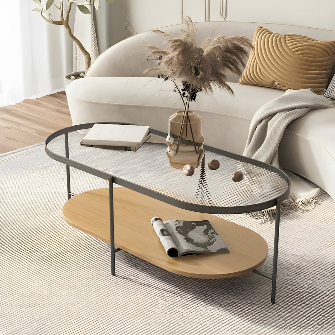 An elliptical coffee table with a clear glass top, a light wood lower shelf, and dark metal legs in a living room setting.