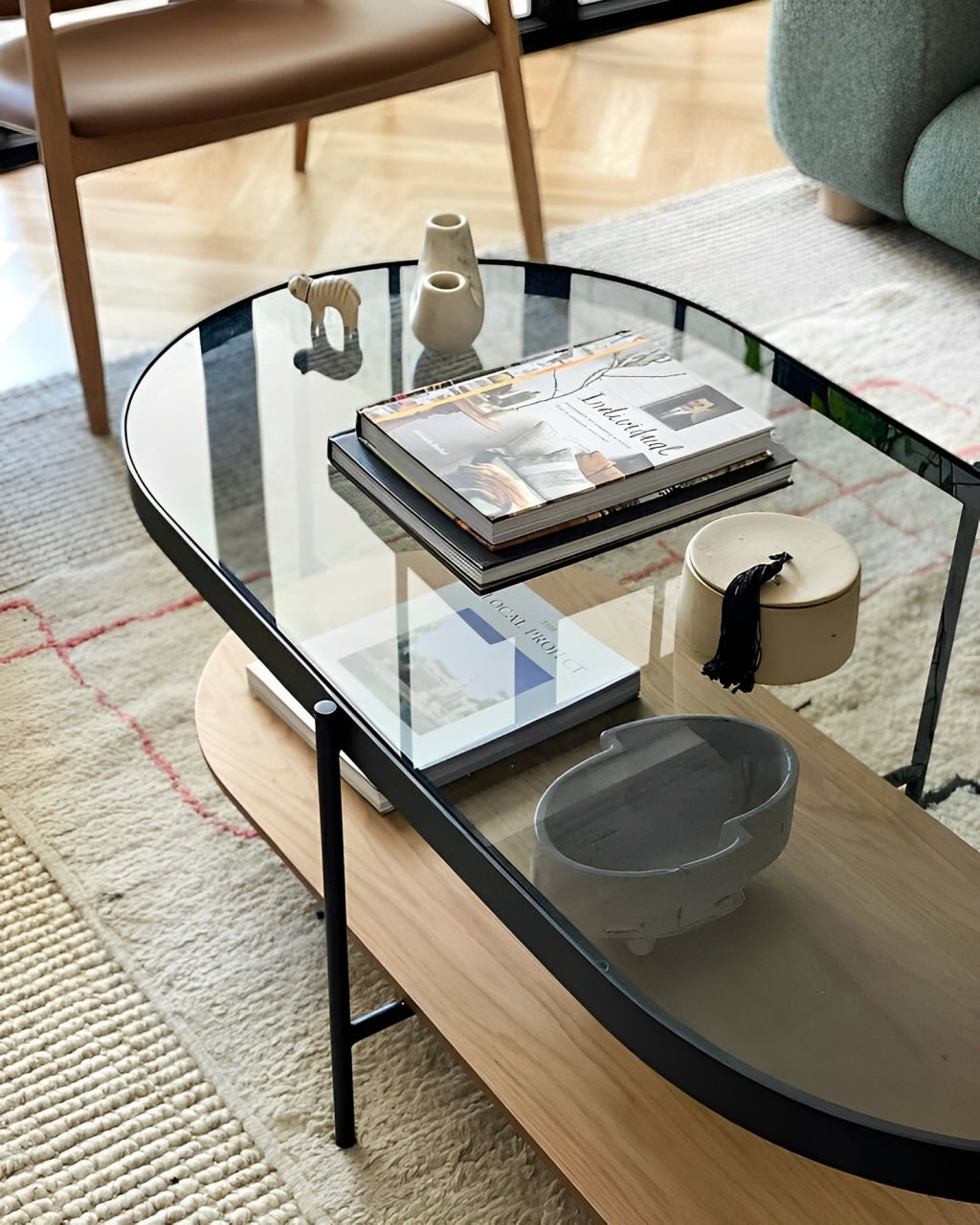 An elliptical coffee table with a clear glass top, a light wood lower shelf, and dark metal legs in a living room setting.