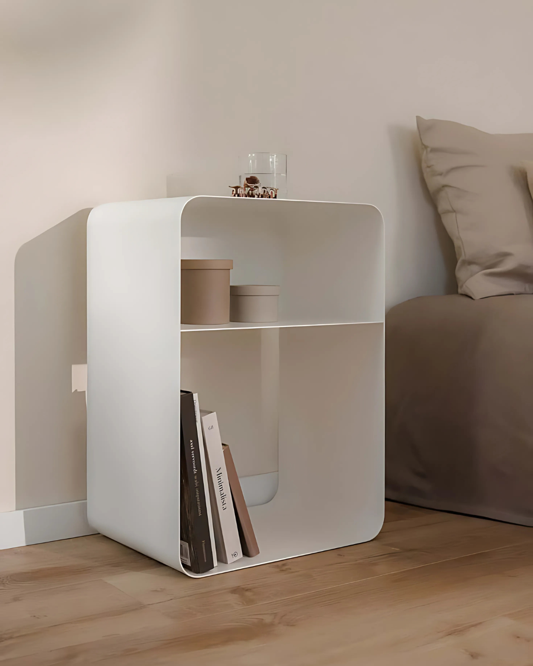 A clean and contemporary bedroom scene featuring a white, two-tiered cubic side table next to a bed. The table holds books and a glass, emphasizing its functionality and minimalist design.