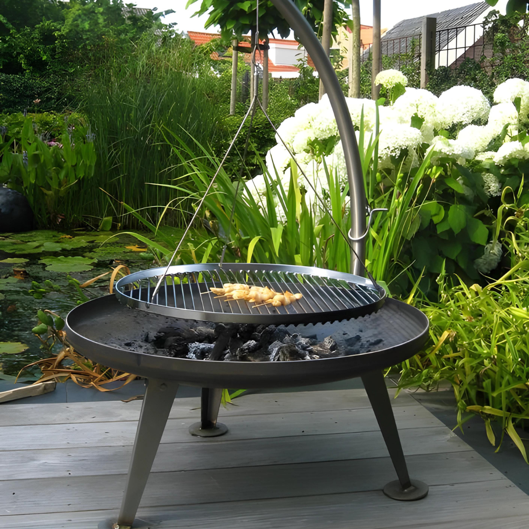 A dark metal fire pit on a wooden deck, featuring a round grill suspended by a metal arm over hot coals, with food cooking.