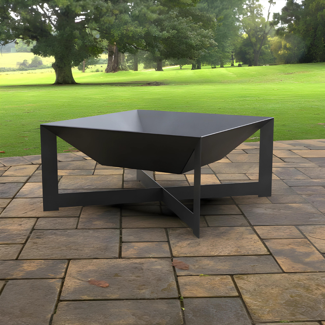 A dark gray, square metal fire pit with a bowl-shaped basin, resting on a stone-tiled patio with a green lawn and trees in the background.