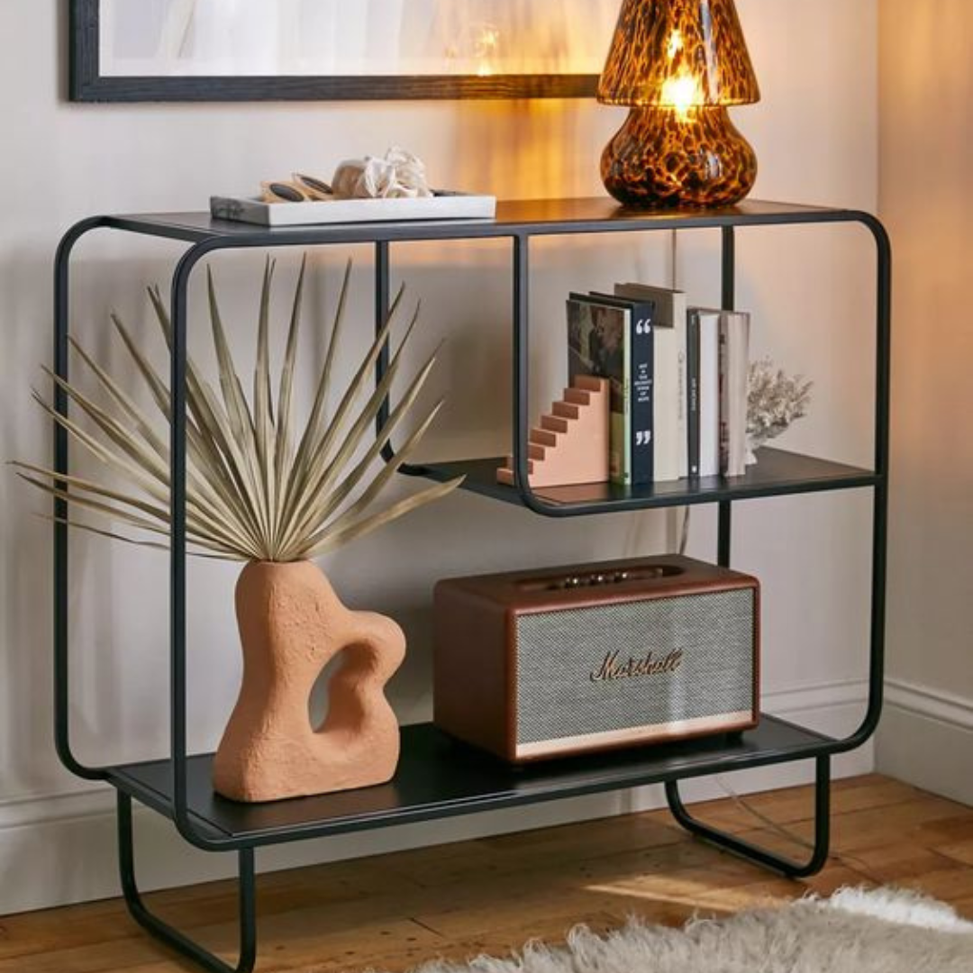 The Alana console, a black metal table with shelves, showing a vase, books, and a speaker.