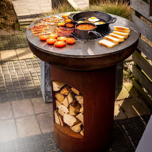 Bonfeu BonBiza Open fire pit grill set on a stone surface, with a cast iron pan of beans and fried eggs, grilled bacon, sausages, tomato halves, and toasted bread slices on the hot plate. Wood is visible in the storage area below.