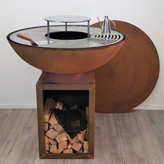 A rusty, bowl-shaped metal fire pit with a flat grilling surface and a center hole. It sits on a rectangular, open-sided stand filled with firewood. A large, round rusty metal cover leans against the wall behind it.