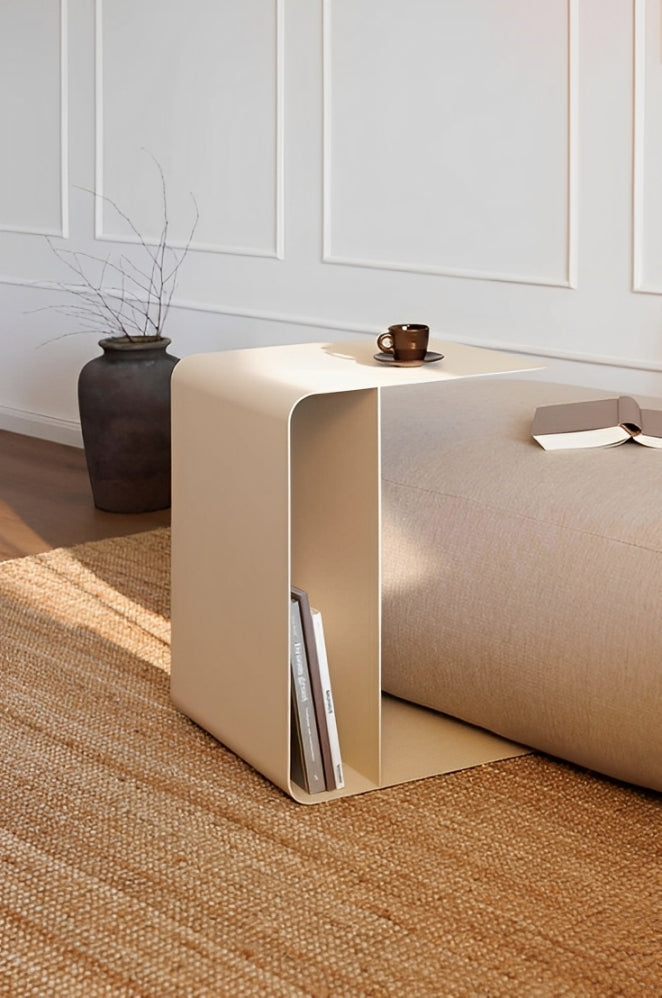 Beige metal C-shaped side table with books in bottom shelf, small vase and glass on top, in modern living room setting.