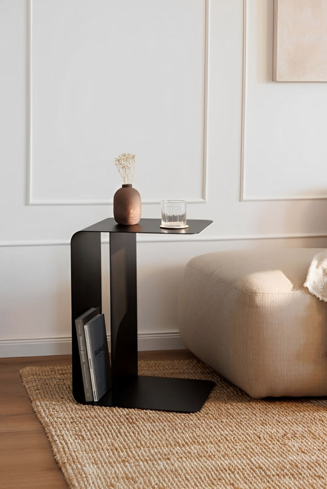 Black metal C-shaped side table with books in bottom shelf, small vase and glass on top, in modern living room setting.